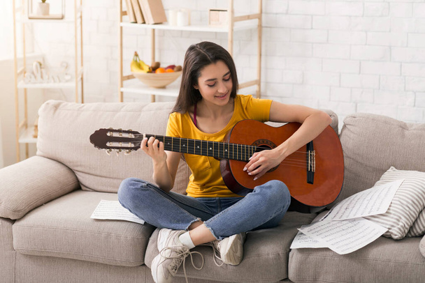 Μείνε σπίτι, διασκέδασε. Υπέροχο μουσικό κορίτσι με νότες που παίζει ακουστική κιθάρα σε άνετο καναπέ σε εσωτερικούς χώρους - Φωτογραφία, εικόνα