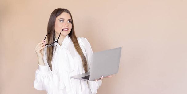 The girl uses a laptop, she 's business, wearing glasses and a white shirt. A young woman with a computer on a beige one-ton background. - Foto, Imagen
