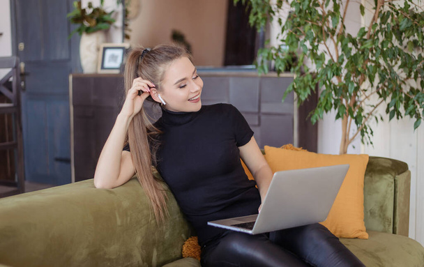 The girl talks to a colleague and friend in an online laptop program. A young woman with wireless headphones works at home with a computer. Online conference. - Фото, изображение