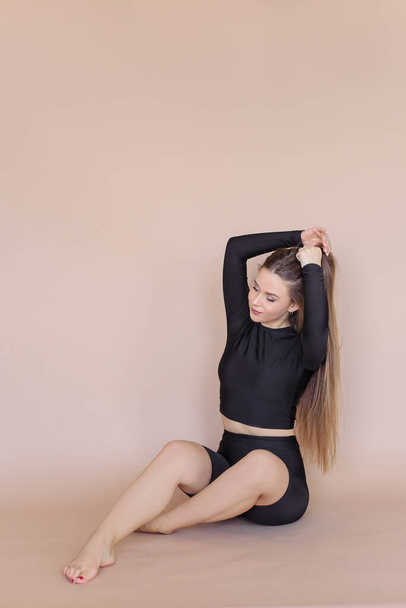 A girl in a black sports suit sits on the floor on a beige background. A beautiful young woman with long hair. - Fotoğraf, Görsel