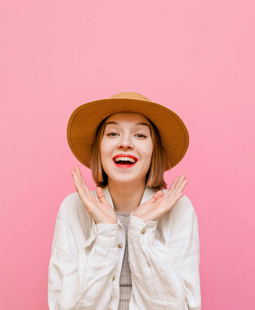 Portrait of a joyful girl in a hat and light summer clothes stands on a pastel pink background and rejoices with palms raised, looks in camera and shouts. Isolated. Vertical - Foto, Imagem