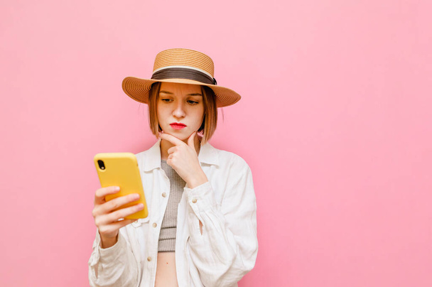Thoughtful girl in hat and shirt on top stands on pink background with smartphone in hand, looks into the screen and thinks. Pensive lady with smartphone looking on smartphone. Copy space - Photo, Image
