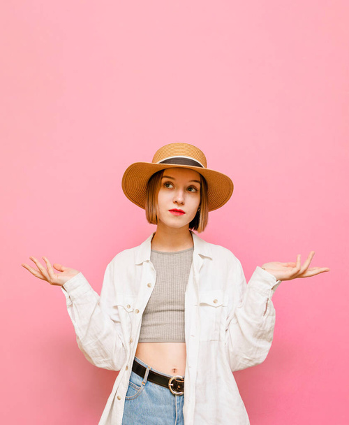 Grappige vrouw in een hoed kijken verbaasd met opgeheven armen van de verbijsterde handen op een pastel roze achtergrond. Portret van verbaasd meisje in lichte kleding spreidt haar handen, geïsoleerd. - Foto, afbeelding