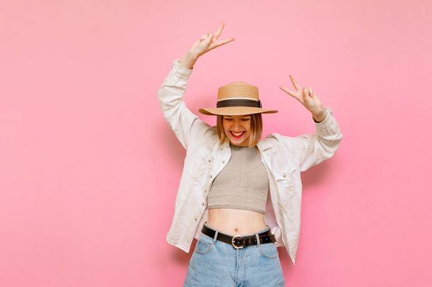 Happy woman in hat and summer clothes rejoices with raised hands on pink background, looks down at copy space and shows a gesture of peace.Joyful lady tourist isolated on pink background. - Foto, afbeelding
