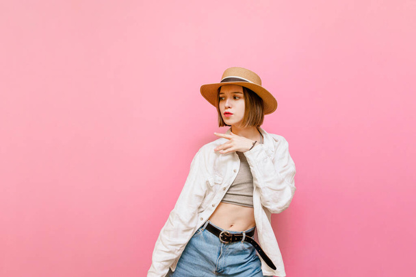 Pretty cheerful girl in light clothing and hat dancing on pink background. Attractive tourist girl dancing and looking aside, isolated on pastel pink background.Copy space - Фото, изображение