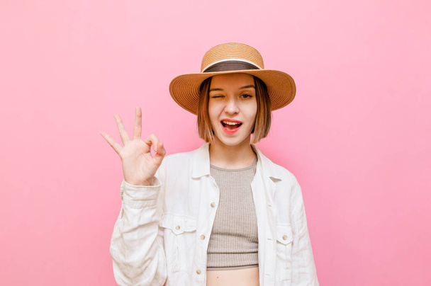 Close-up portret van een grappig meisje in een hoed en lichte kleding op een roze achtergrond, kijken in de camera met een glimlach op haar gezicht en tonen een OK gebaar en knipogen. Kopieerruimte - Foto, afbeelding