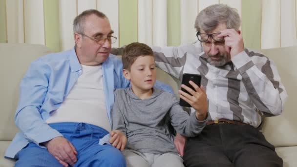Twee grootvaders met een kleinzoon op de bank kijken naar de smartphone die de kleinzoon hen laat zien. - Video