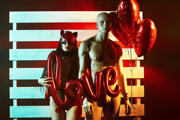 Sexy woman wearing a black cat mask standing hugging mannequin and holding heart-shaped balloons and a love sign and looks very sensually - Photo, image
