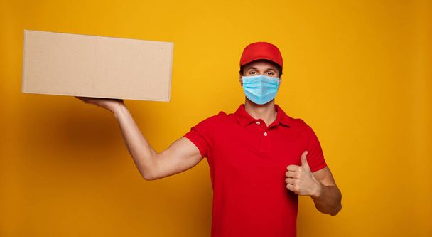 Young delivery man in medical safety mask holding and carrying a card box isolated on yellow background. Buy food online in the quarantine period. - Photo, image