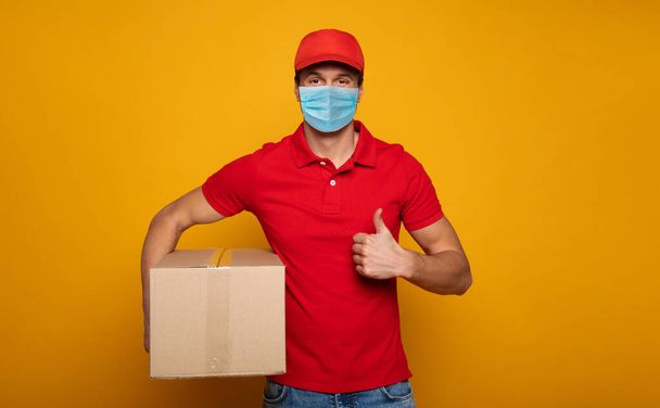 Young delivery man in medical safety mask holding and carrying a card box isolated on yellow background. Buy food online in the quarantine period. - Photo, image