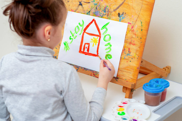 Niño está dibujando casa roja por acuarelas con una frase escrita sobre la casa Stay Home en el caballete. Concepto Stay Home
. - Foto, Imagen