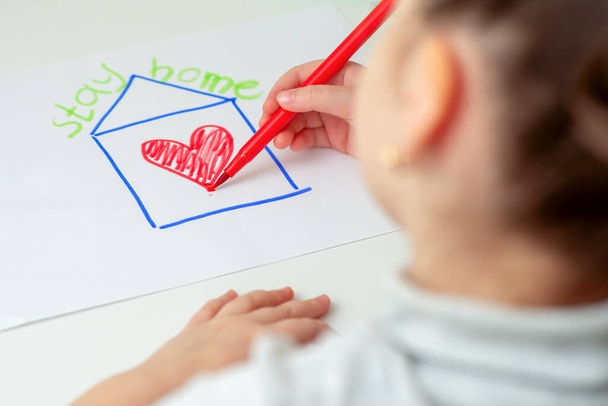 Evde Kal konsepti. Çocuk, kırmızı kalpli bir evi kağıdın üzerine çiziyor. Evin üzerinde yazılı bir ifade var. Evde, sehpanın üzerinde kal.. - Fotoğraf, Görsel