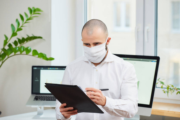 Muž v lékařské masce obličeje proti koronaviru (COVID-19). Kancelářský pracovník si dělá poznámky na svém pracovišti s počítači a zelenými rostlinami v pozadí. Karanténa koronaviru - Fotografie, Obrázek