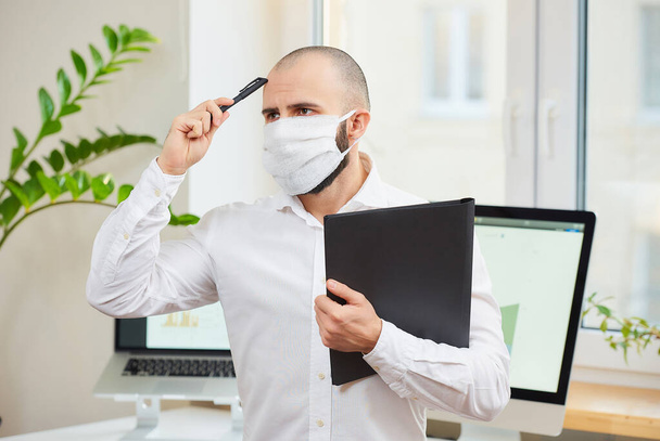 Ein Mann mit einer medizinischen Gesichtsmaske gegen das Coronavirus (COVID-19). Ein Ingenieur kratzt sich mit einem Stift an seinem Arbeitsplatz mit Computern und einer grünen Pflanze im Hintergrund den Kopf. Coronavirus-Quarantäne. - Foto, Bild