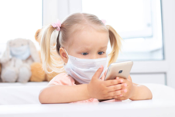 girl in a protective medical mask using a mobile phone, a smartphone for video calls, talks with relatives, a girl sits at home, online computer webcam, making a video call. - Φωτογραφία, εικόνα
