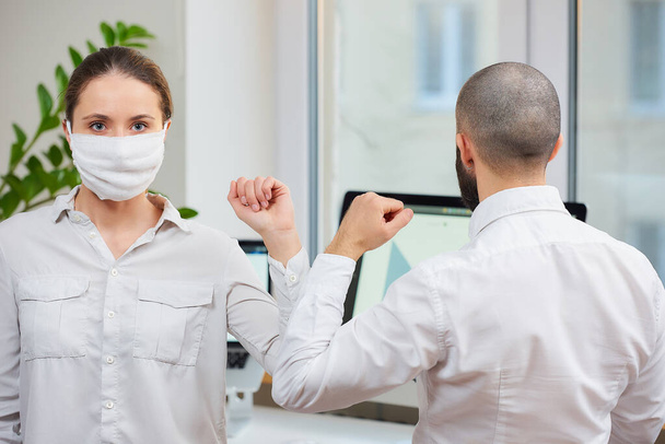 Coronavirus 'un (2019-nCoV) yayılmasını önlemek için dirsek seği selamı. Tıbbi maskeli bir kadın ve bir adam ofiste çıplak elleriyle buluşuyor. Sarılmak ya da tokalaşmak yerine, dirseklerini vururlar. - Fotoğraf, Görsel