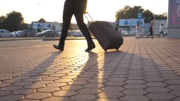 Voeten van succesvolle zakenman lopen vanaf het vliegveld met zijn bagage op straat. Poten van de jonge manager die bij zonsondergang naar de parkeerplaats gaat en een koffer op wielen trekt. Slow motion zijaanzicht - Video
