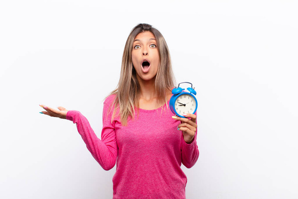 young pretty woman looking surprised and shocked, with jaw dropped holding an object with an open hand on the side holding an alarm clock. - 写真・画像