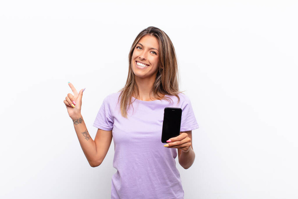 young pretty woman looking excited and surprised pointing to the side and upwards to copy space holding a smart telephone - Фото, зображення