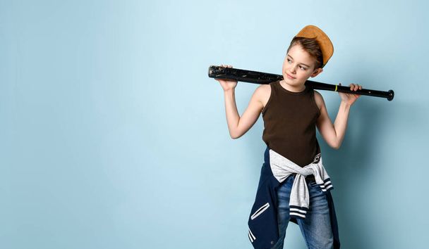 Sly little schoolboy stands near the wall looking aside holding baseball bat on shoulders. Three quarter length portrait isolated on blue background - Foto, Imagem