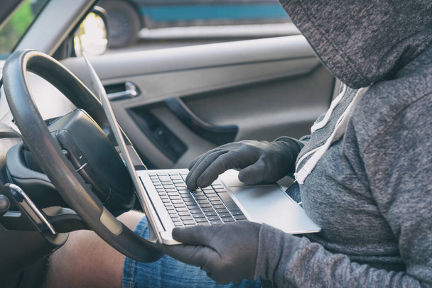 フード付き泥棒は、ラップトップで車のセキュリティシステムを破壊しようとします。現代的な車のコンセプトをハッキング - 写真・画像