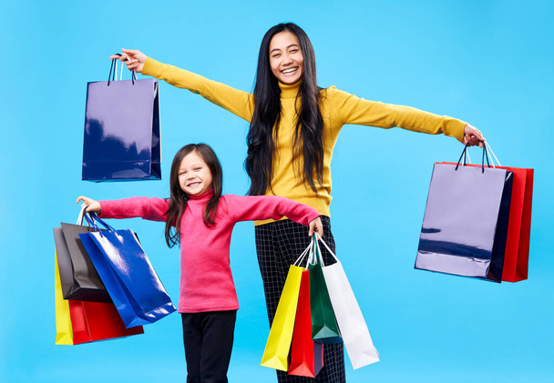 Happy mother with adorable little daughter holding shopping bags enjoy their purchase on blue background. - Photo, Image