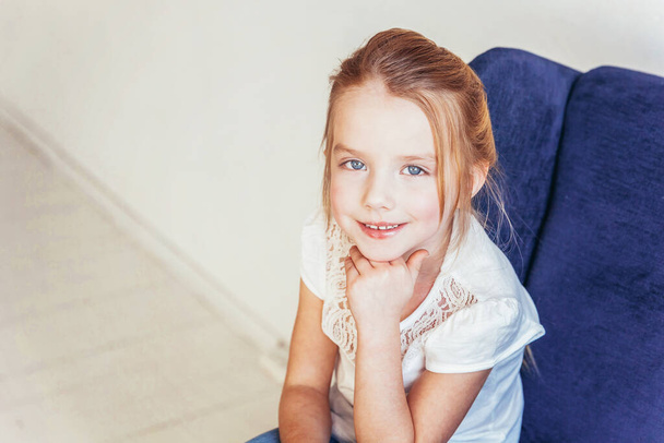 Оставайся дома. Милая маленькая девочка в джинсах и белой футболке сидит на современном уютном синем стуле, расслабляясь в белой яркой гостиной в помещении. Детство школьники молодежь расслабиться концепция
. - Фото, изображение