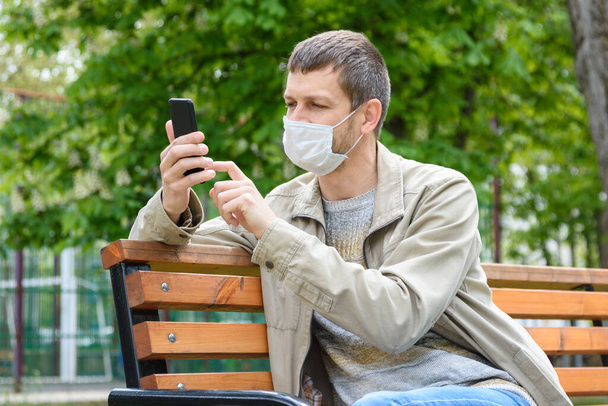 Ένας άντρας με ιατρική μάσκα και smartphone κάθεται σε ένα παγκάκι στο πάρκο. - Φωτογραφία, εικόνα