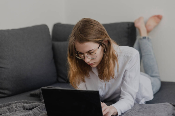 Attraktive junge Frau, die von zu Hause aus arbeitet - Unternehmerin, die auf einem Sofa mit Laptop sitzt und das Handy von zu Hause aus checkt. Fernarbeit - Foto, Bild