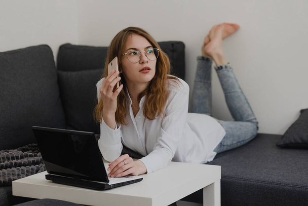 Attraktive junge Frau, die von zu Hause aus arbeitet - Unternehmerin, die auf einem Sofa mit Laptop sitzt und das Handy von zu Hause aus checkt. Fernarbeit - Foto, Bild