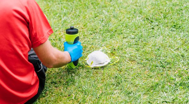 コピースペースと美しい緑の芝生の公園でコロナウイルスの拡散を防ぐために保護マスク付きウォーターボトルを保持スポーツマン. - 写真・画像