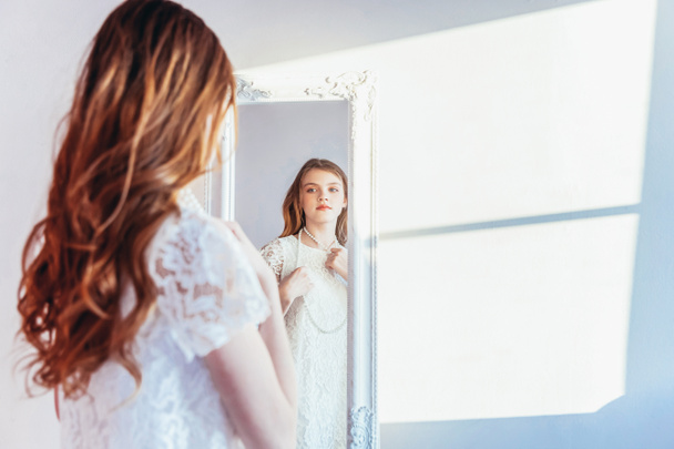 Beauty makeup morning rutine love yourself concept. Young teenage girl looking at reflection in mirror. Young positive woman wearing white dress posing in bright light room against white wall. - Foto, immagini