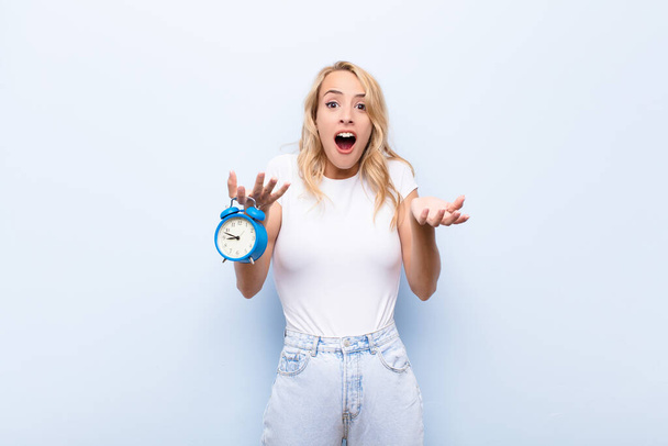 young blonde woman open-mouthed and amazed, shocked and astonished with an unbelievable surprise holding a clock - Fotó, kép