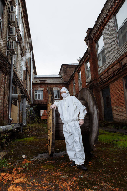 mężczyzna w kombinezonie ochronnym, białej masce medycznej i rękawiczkach lateksowych podczas pandemii koronawirusowej COVID-19 w ruinach miasta - Zdjęcie, obraz