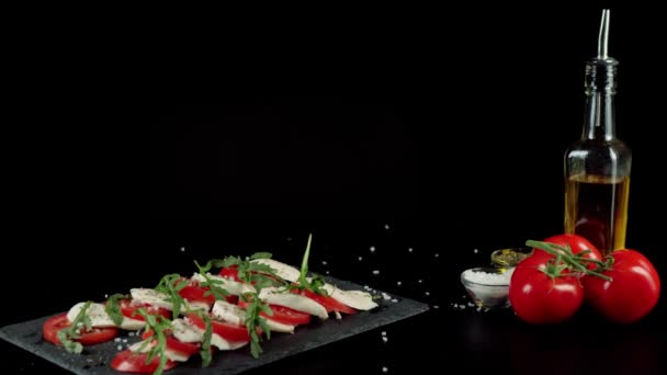 Kamera włącza gotową sałatkę caprese. Świeże soczyste pomidory, ser mozzarella i liście rukoli na czarnej tablicy z ziołami śródziemnomorskimi i solą morską. Środkowy strzał. 4K - Materiał filmowy, wideo
