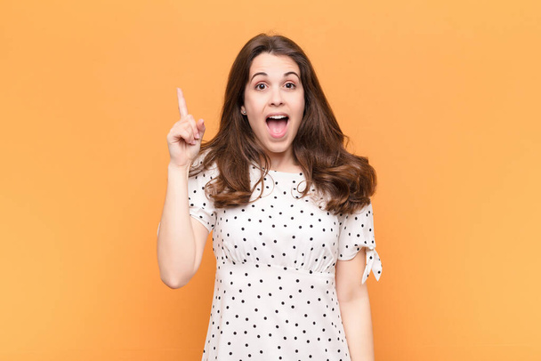 young pretty woman feeling like a happy and excited genius after realizing an idea, cheerfully raising finger, eureka! against orange wall - Fotoğraf, Görsel