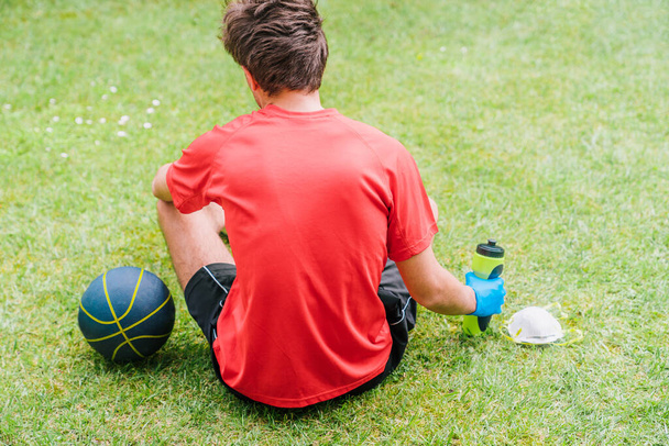 Αθλητικό αγόρι που ασκεί υπαίθρια αθλήματα με προστατευτικά μέτρα για την πρόληψη της εξάπλωσης του coronavirus. Πρόσωπο με ένα μπάσκετ, μια μάσκα και ένα μπουκάλι νερό κάθεται στο γρασίδι του πάρκου. - Φωτογραφία, εικόνα
