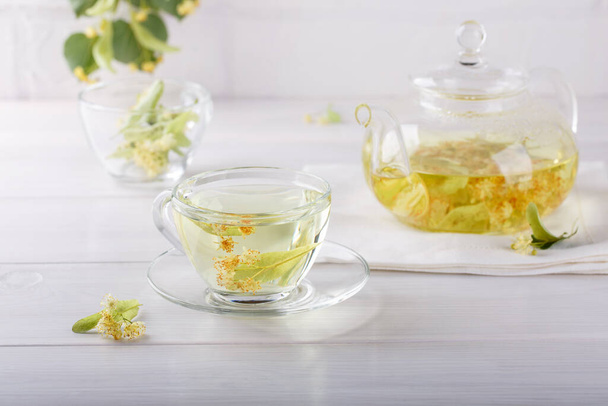 Linden tea in a transparent tea pot.  Glass cup and tea pot with herbal lime tree tea on white background. Medicinal plant, flowers used for herbal teas and tinctures. - Photo, Image