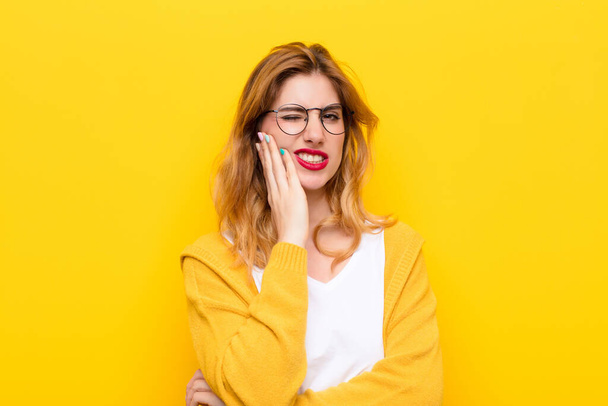young pretty blonde woman holding cheek and suffering painful toothache, feeling ill, miserable and unhappy, looking for a dentist against yellow wall - Photo, image