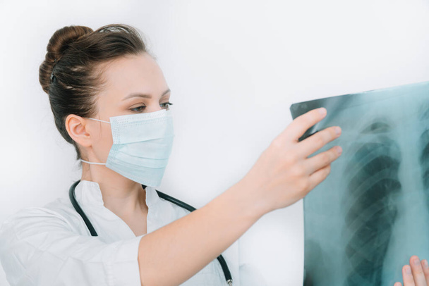 Young caucasian doctor in protective mask examine x-ray of lungs, holding it in hands indoors. Specialist hold transparent image of chest in arms, and carefully researches it for coronavirus - Φωτογραφία, εικόνα