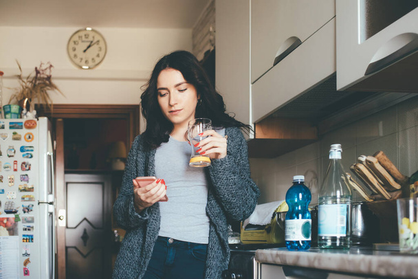 Jovem bela mulher do Oriente Médio em casa cozinha usando suco de bebida smartphone rotina matinal, tecnologia 5g, conceito de folga
 - Foto, Imagem
