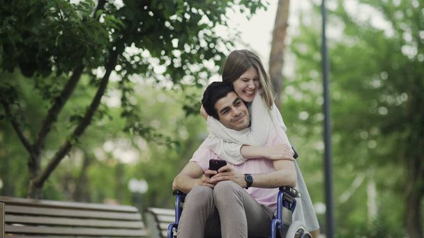 Ο νεαρός ανάπηρος είναι ευχαριστημένος με τις αγκαλιές της γυναίκας που αγαπάει. Πηγαίνουν βόλτα στο πάρκο. - Φωτογραφία, εικόνα
