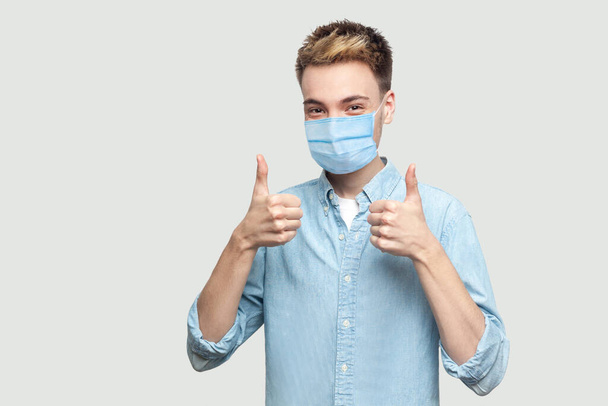 Portrait of happy satisfied successful handsome young man with surgical medical mask in light blue shirt standing, thumbs up and looking at camera. indoor studio shot on grey background copy space. - Foto, imagen