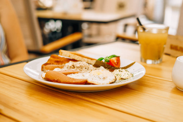 Delicious breakfast in the cafe: scrambled eggs and a glass of juice. Scrambled eggs with sausages, tomatoes and cheese. Scrambled eggs with ham and toast on a white round plate - Fotó, kép
