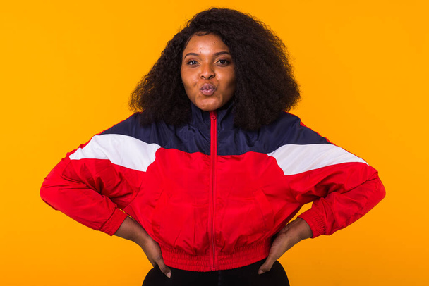Funny curly african american girl dressed in the red sports jacket in the studio on yellow background. Fashion and people concept. - 写真・画像