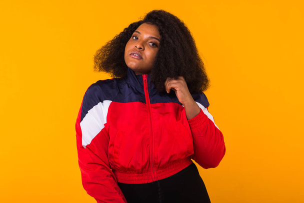 Divertente ragazza afroamericana riccia vestita con la giacca sportiva rossa in studio su sfondo giallo. Moda e concetto di persone
. - Foto, immagini