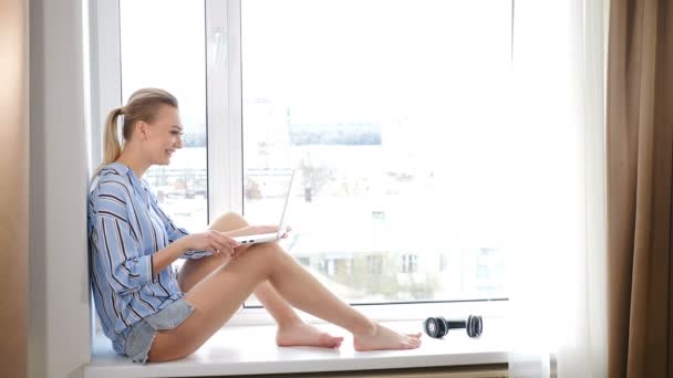 Atrakcyjna młoda kobieta pracująca w domu w kwarantannie z laptopem na kolanach siedząc przy parapecie. Koncepcja pracy online i edukacji. Kobieta freelancer student za pomocą laptopa studiuje online pracy - Materiał filmowy, wideo