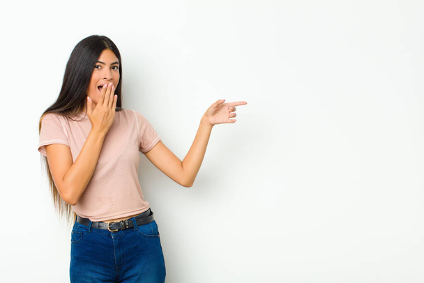 young pretty latin woman feeling happy, shocked and surprised, covering mouth with hand and pointing to lateral copy space against flat wall - Fotó, kép