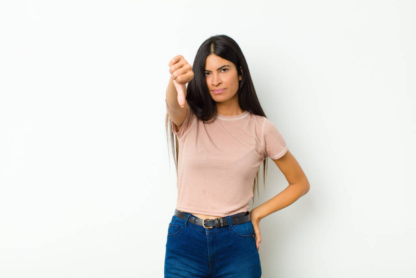 young pretty latin woman feeling cross, angry, annoyed, disappointed or displeased, showing thumbs down with a serious look against flat wall - Photo, image
