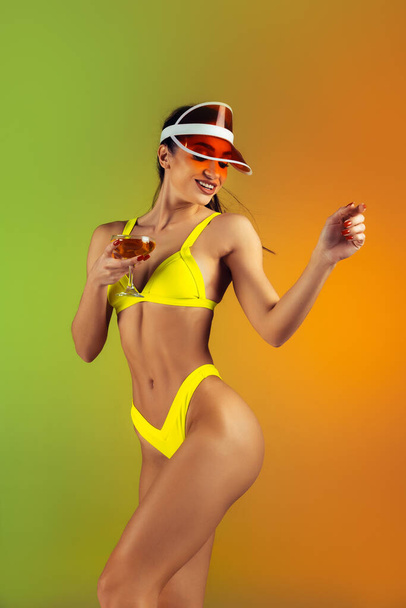 Fashion portrait of young fit and sportive woman with cocktail in stylish yellow luxury swimwear on gradient background. Perfect body ready for summertime. - Φωτογραφία, εικόνα